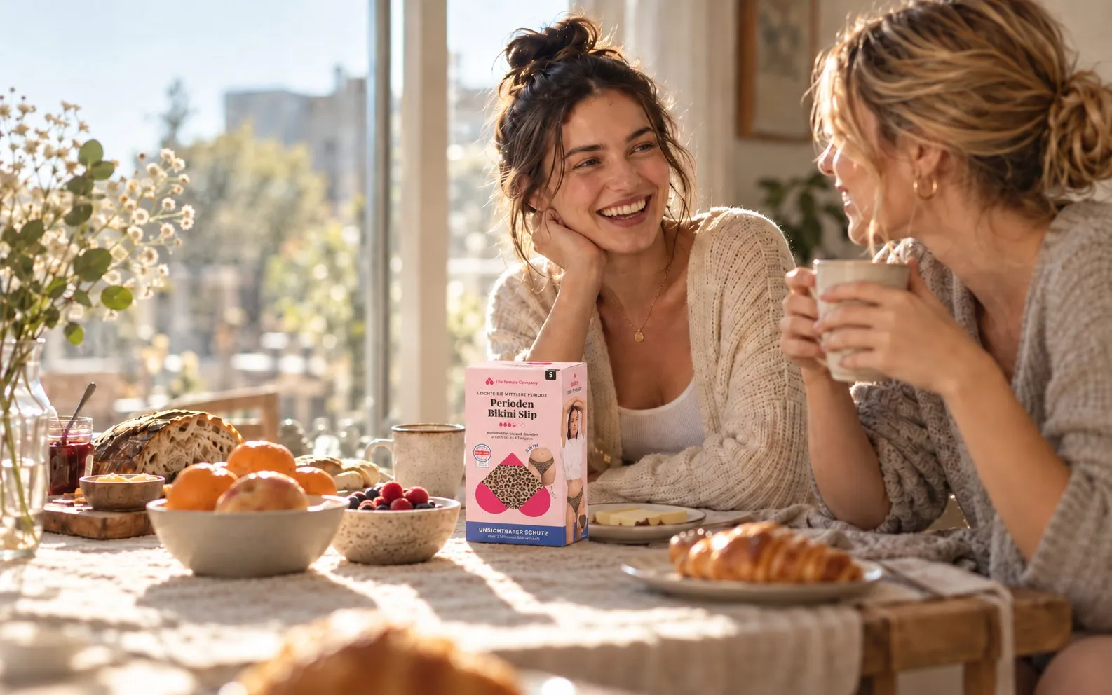 Zwei Frauen lachen am Frühstückstisch, im Vordergrund eine Packung Periodenunterwäsche von The Female Company